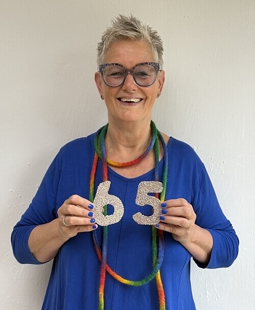 Elly laat lachend zien dat ze 65 jaar geworden is. Ze heeft een 6 en een 5 voor haar borst.